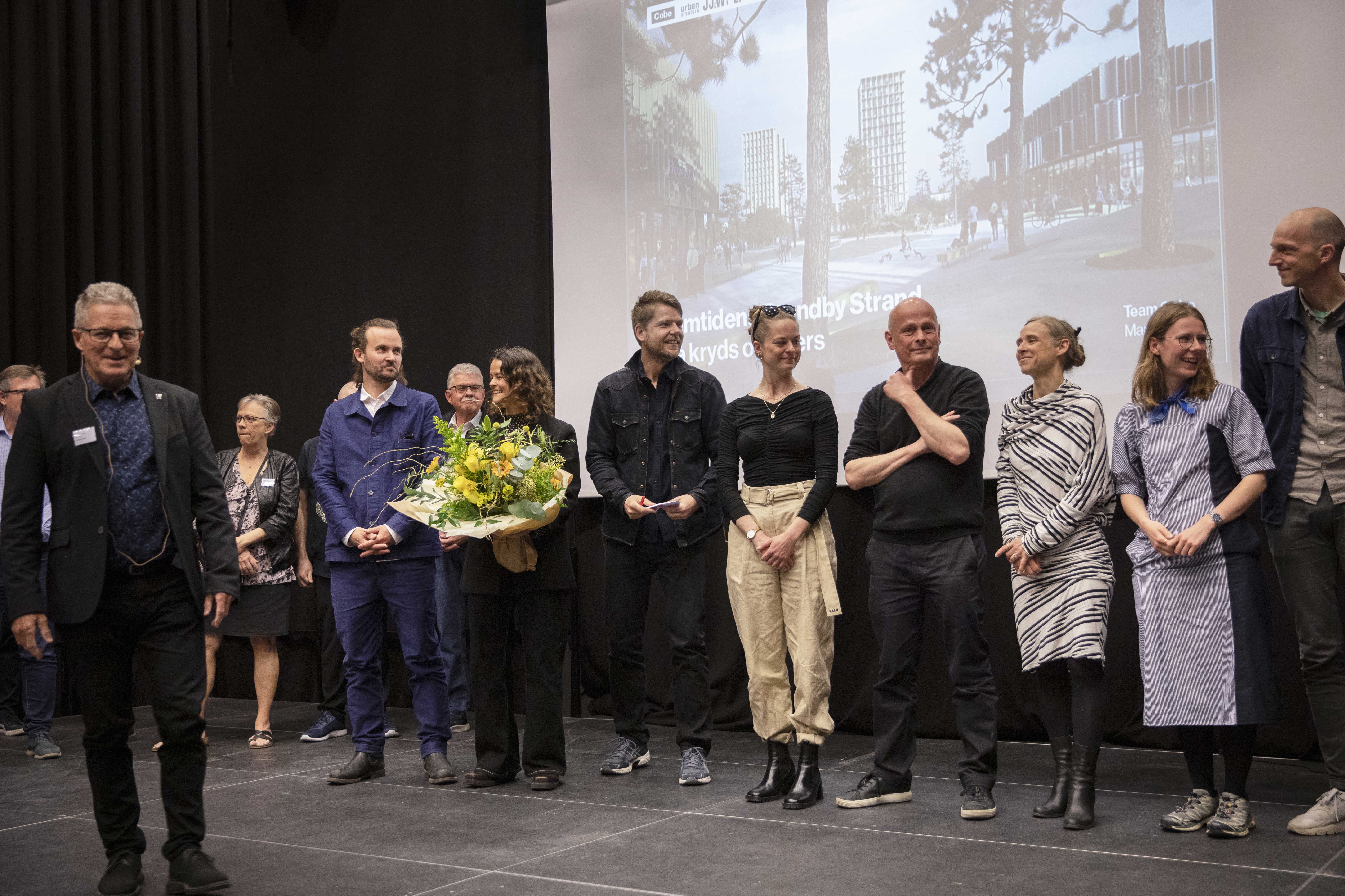 Team Cobe står på scenen i Brønden, en af repræsentanterne for teamet holder en buket med gule blomster. I forggrunden står Borgmester Kent Magelund.